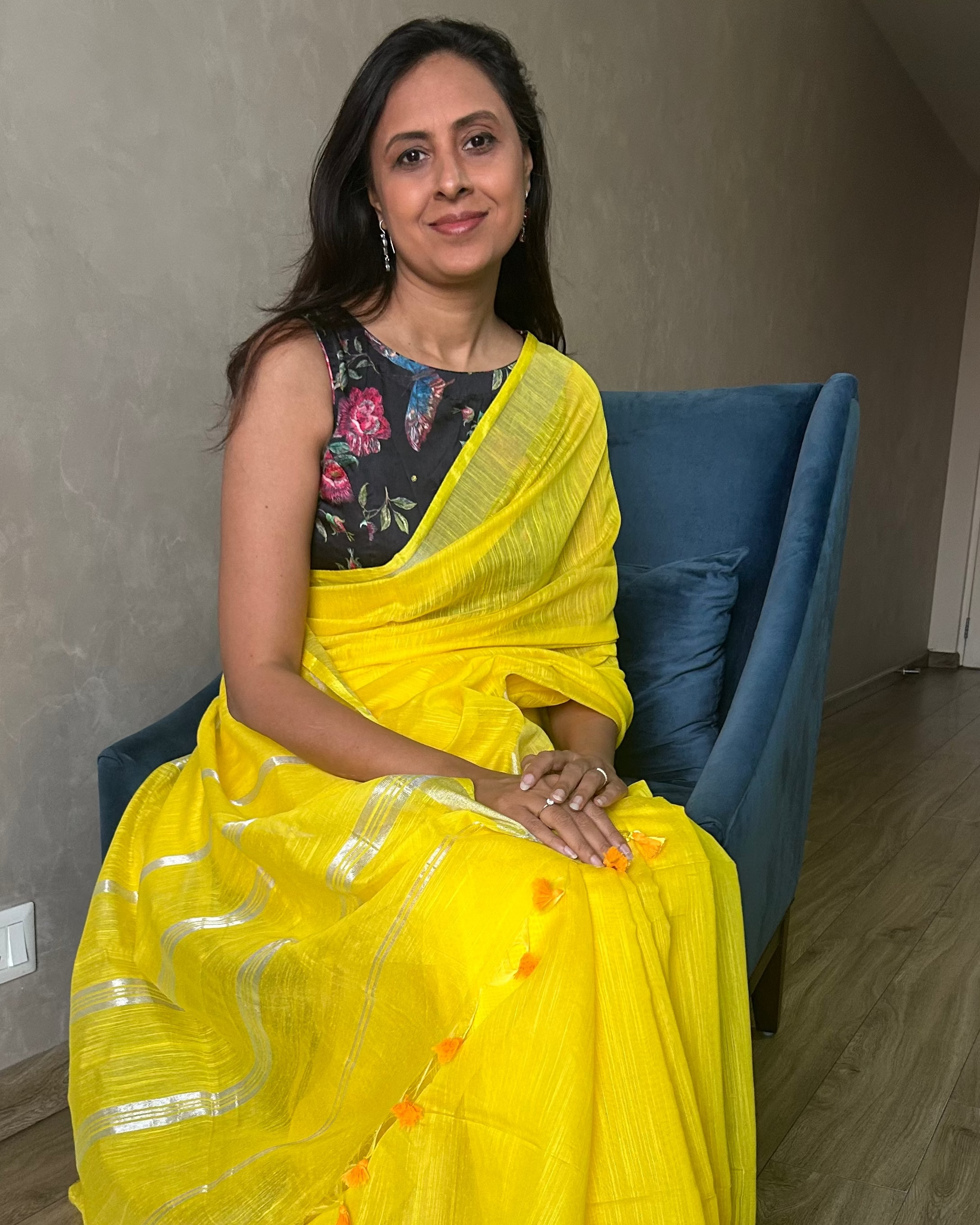 Woman in a yellow saree sitting on a blue chair indoors.